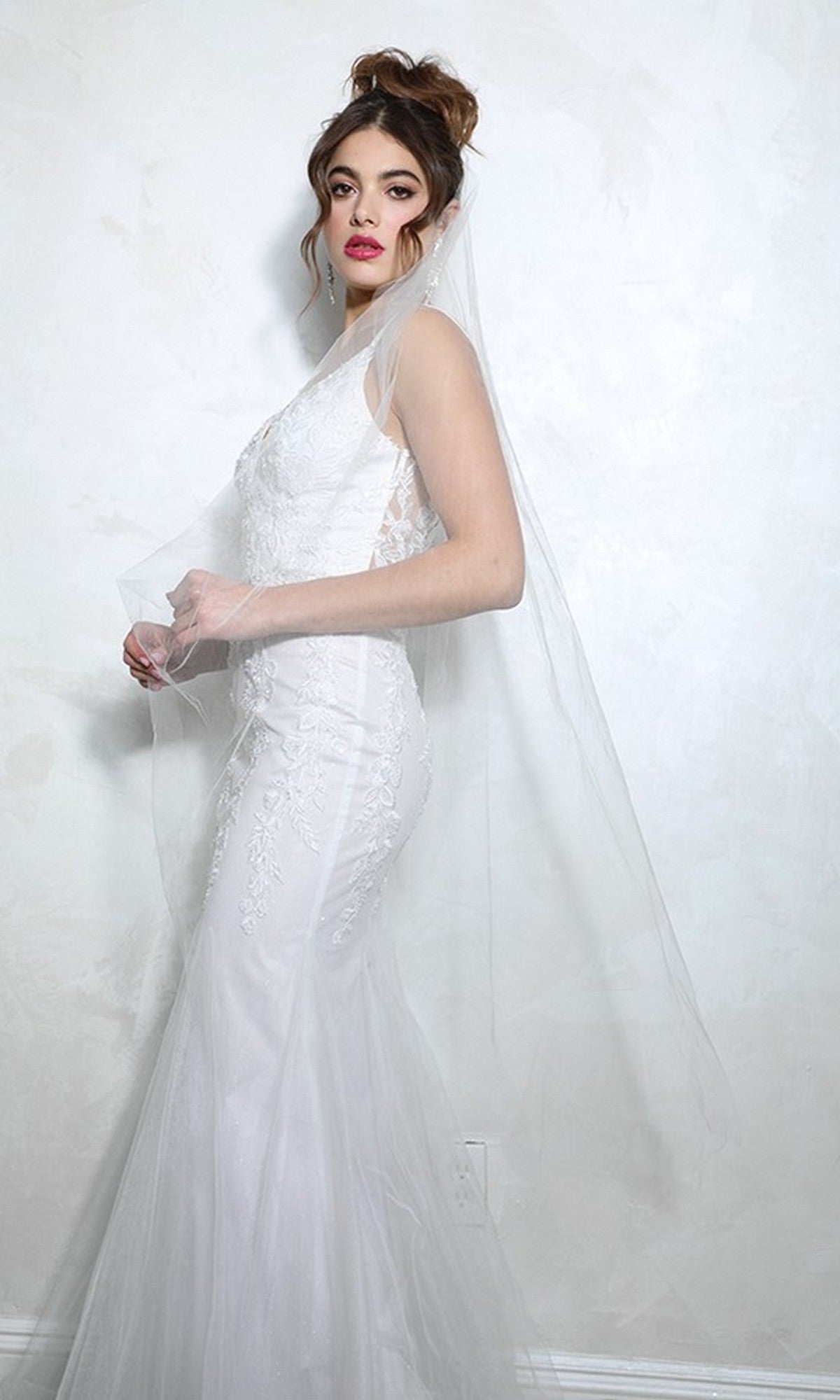 A woman in the off white Eureka 9998 long prom dress stands against a textured white wall, looking to the side.